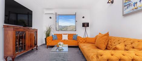 A living room with a large flat-screen TV, a sofa, a coffee table, and a window with a view of hills.