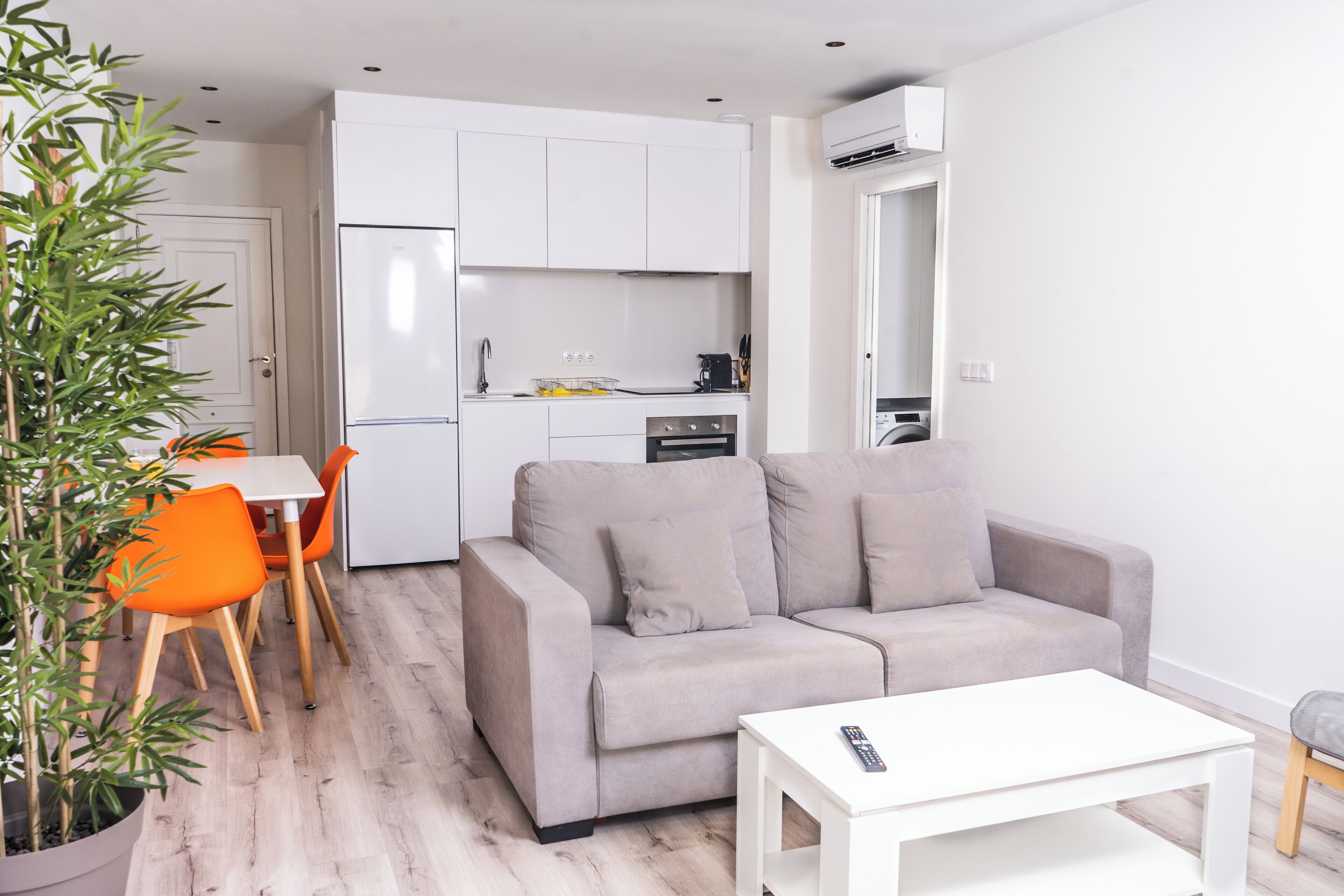 Una sala de estar moderna con un sofá gris, una mesa de centro blanca y sillas naranjas. Se ve una cocina al fondo, con gabinetes blancos y una estufa.