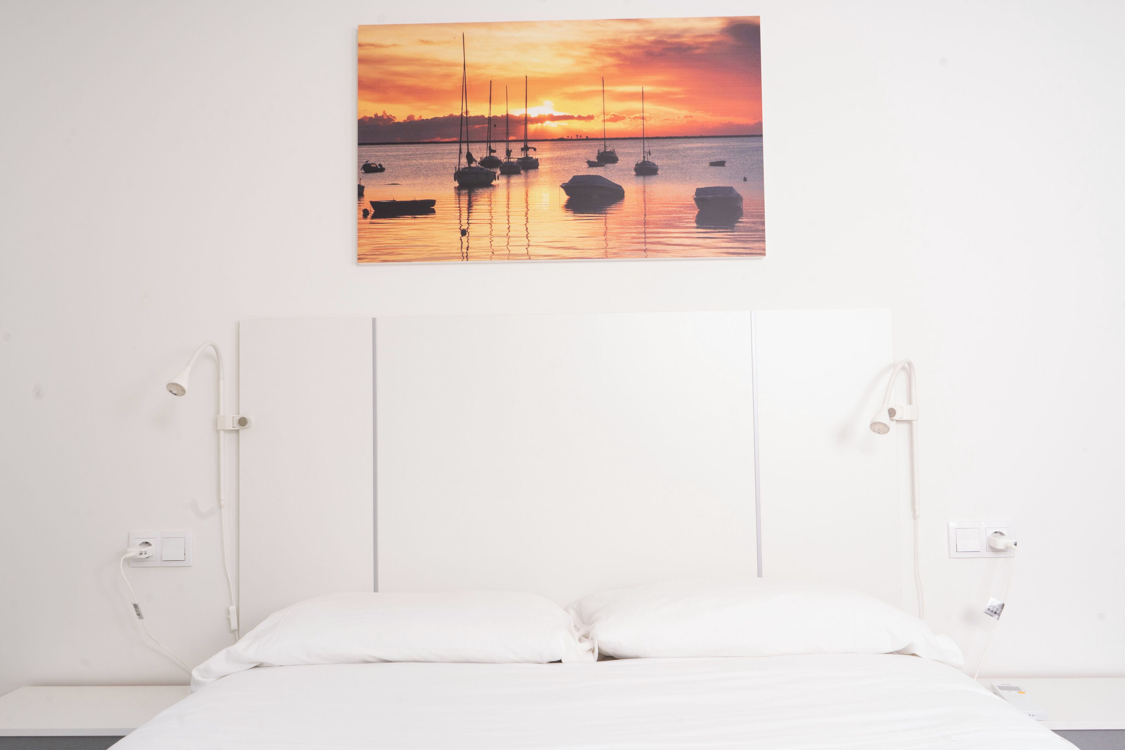 Quarto com cama, cabeceira branca, mesas de cabeceira e uma pintura na parede de barcos ao pôr do sol.