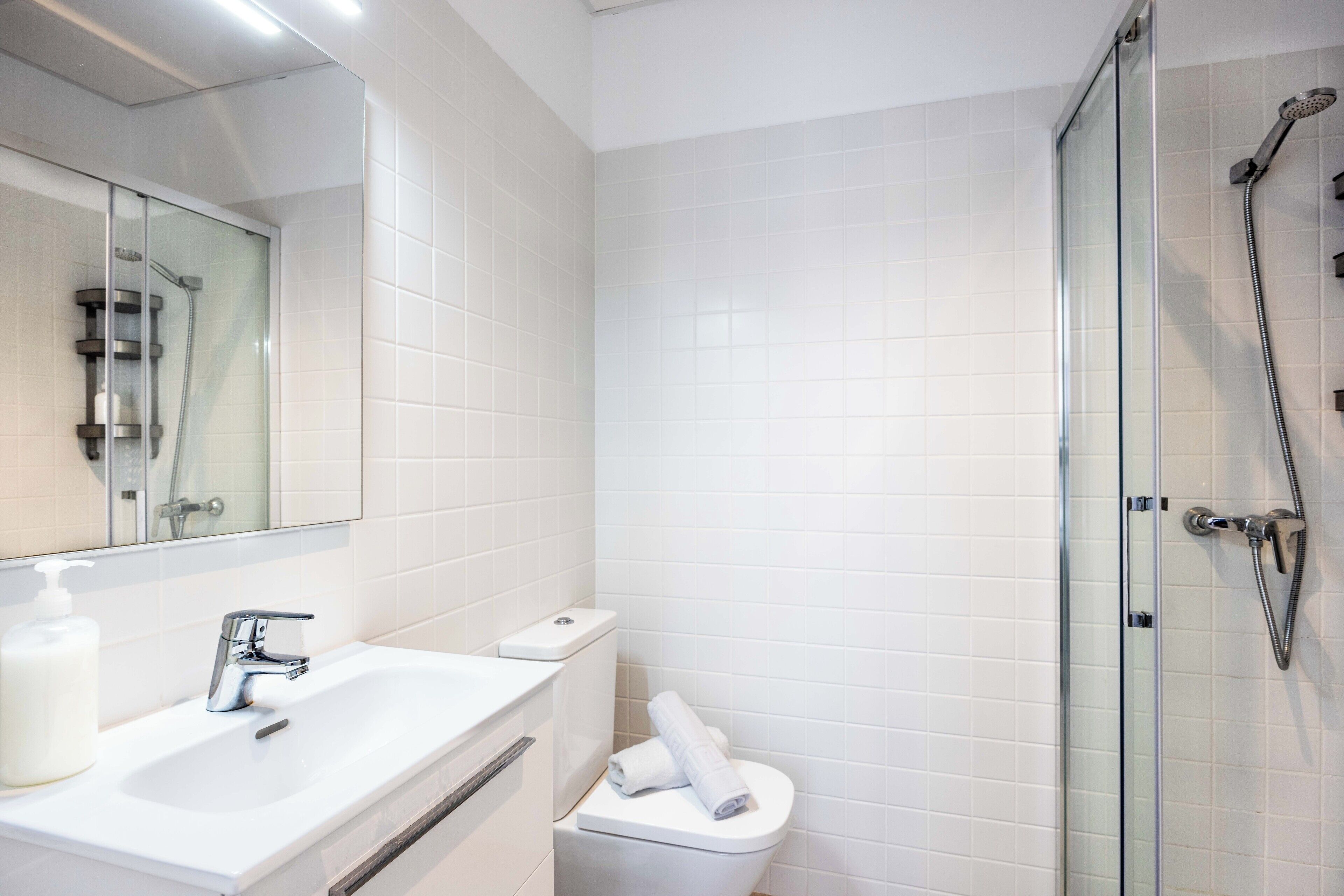 Et moderne badeværelse med et glasbrusekabine, en hvid vask med sæbedispenser og et toilet.
