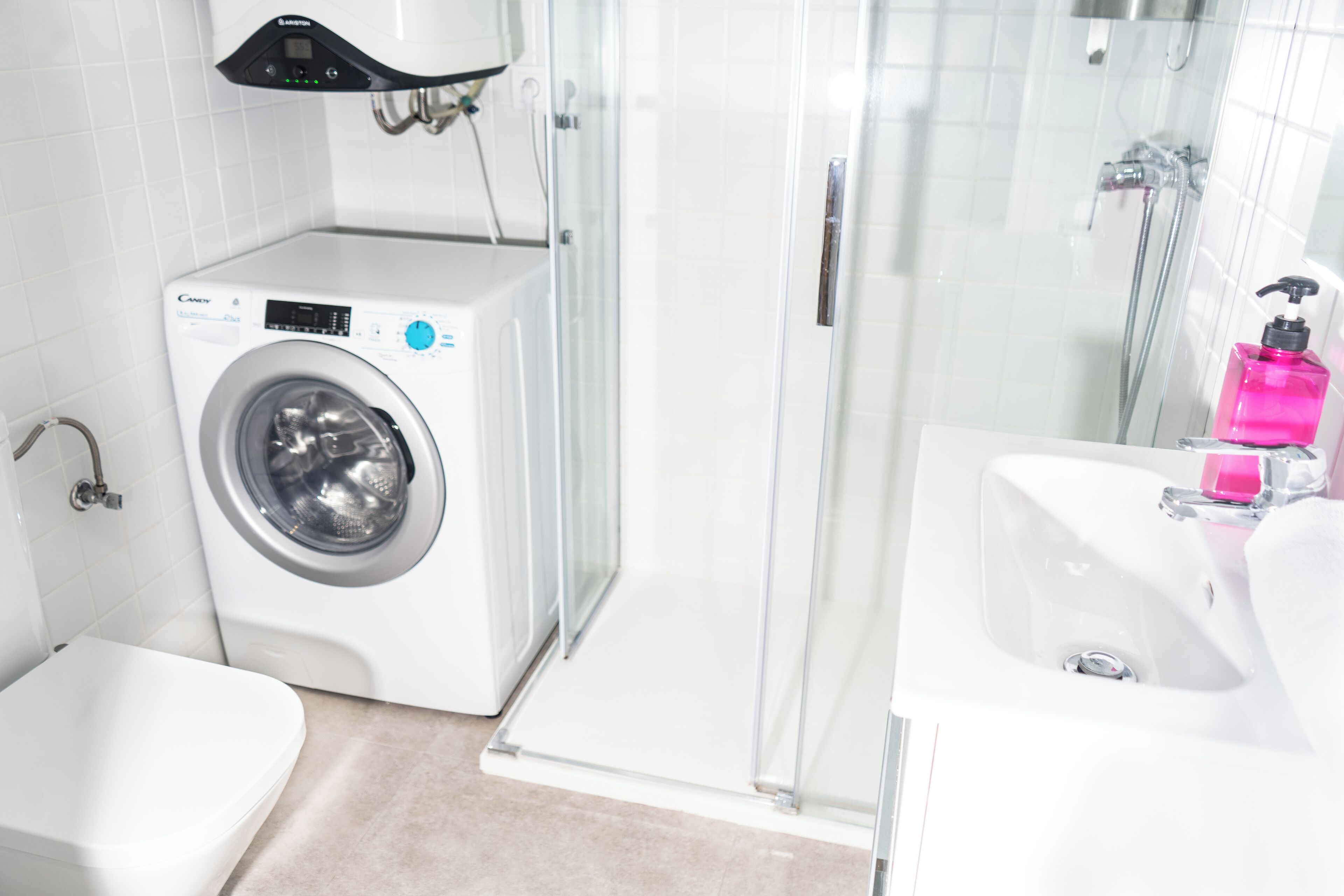 Un baño moderno con lavadora, cabina de ducha de vidrio y lavamanos con dispensador de jabón rosa.