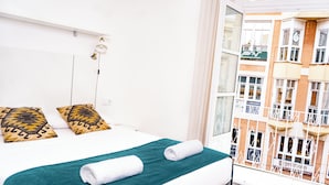 A hotel room with a bed, a green bedspread, two patterned pillows, and a window with a view of a building.