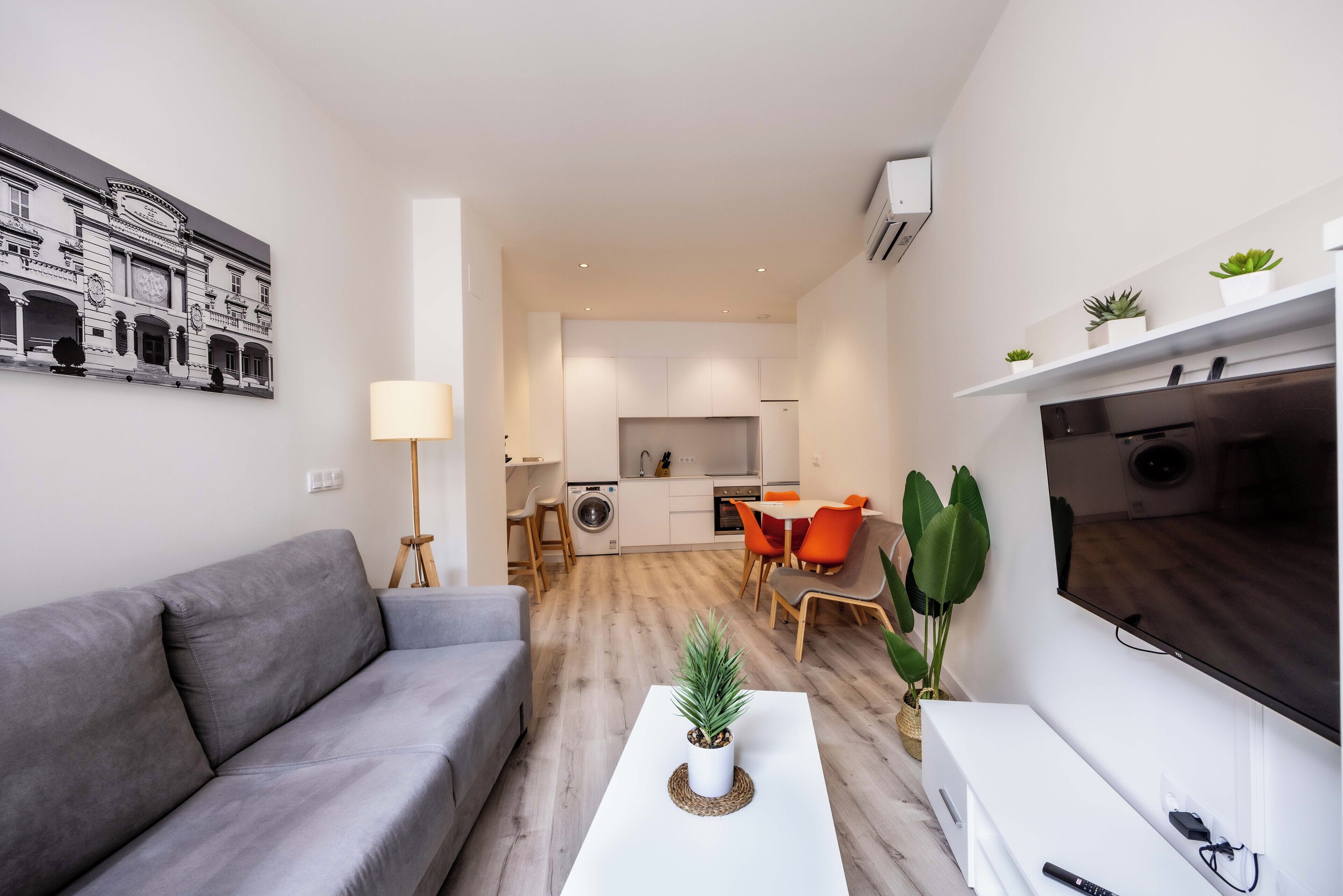 Una sala de estar moderna con un sofá gris, una mesa de centro blanca con una planta, un televisor y una cocina al fondo.