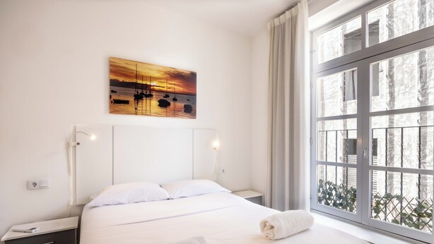 Habitación de hotel con una cama grande, una mesita de noche, una lámpara de pared y una ventana con vistas a zonas verdes.