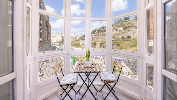 A bay window with a wooden table and two chairs, a potted plant, and a view of a hillside.