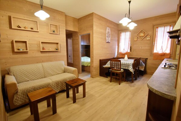 A cozy wooden interior with a sofa, a dining table with chairs, and a small wooden table. Shelves with decorative items are on the walls.