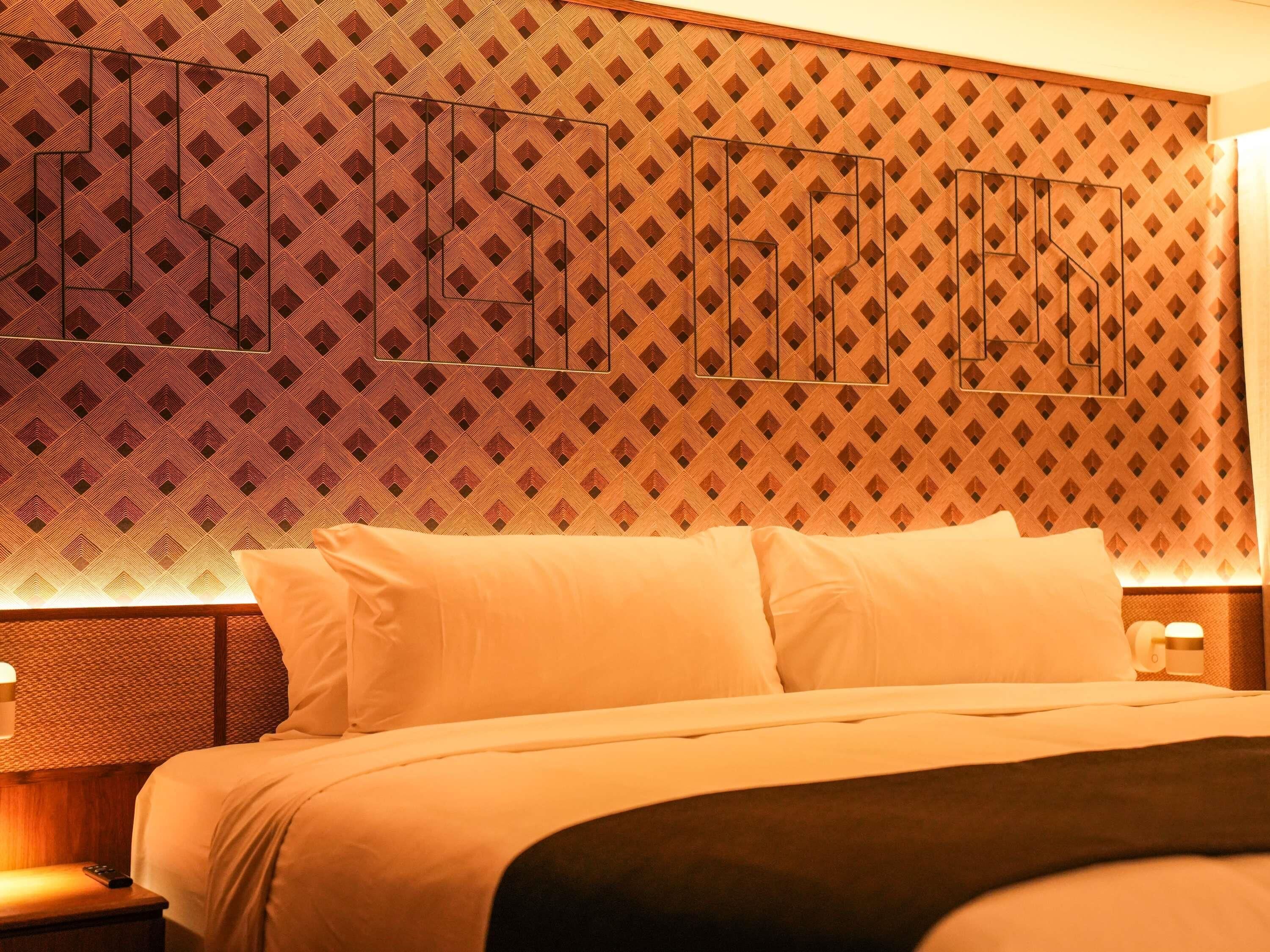 Una camera d'albergo con una testiera a motivi decorativi, un letto con biancheria da letto bianca e un comodino con una lampada.