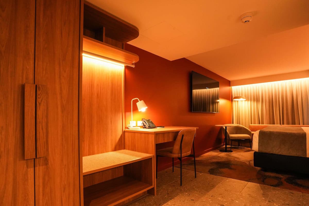 A hotel room with a wooden wardrobe, a desk with a lamp, a chair, a bed, and a window with curtains.