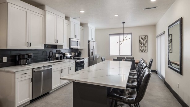 A modern kitchen with white cabinets, stainless steel appliances, a large island with bar stools, and a window providing natural light. - Vizcaya #12 - Moab's Newest Luxury Rental (Moab)