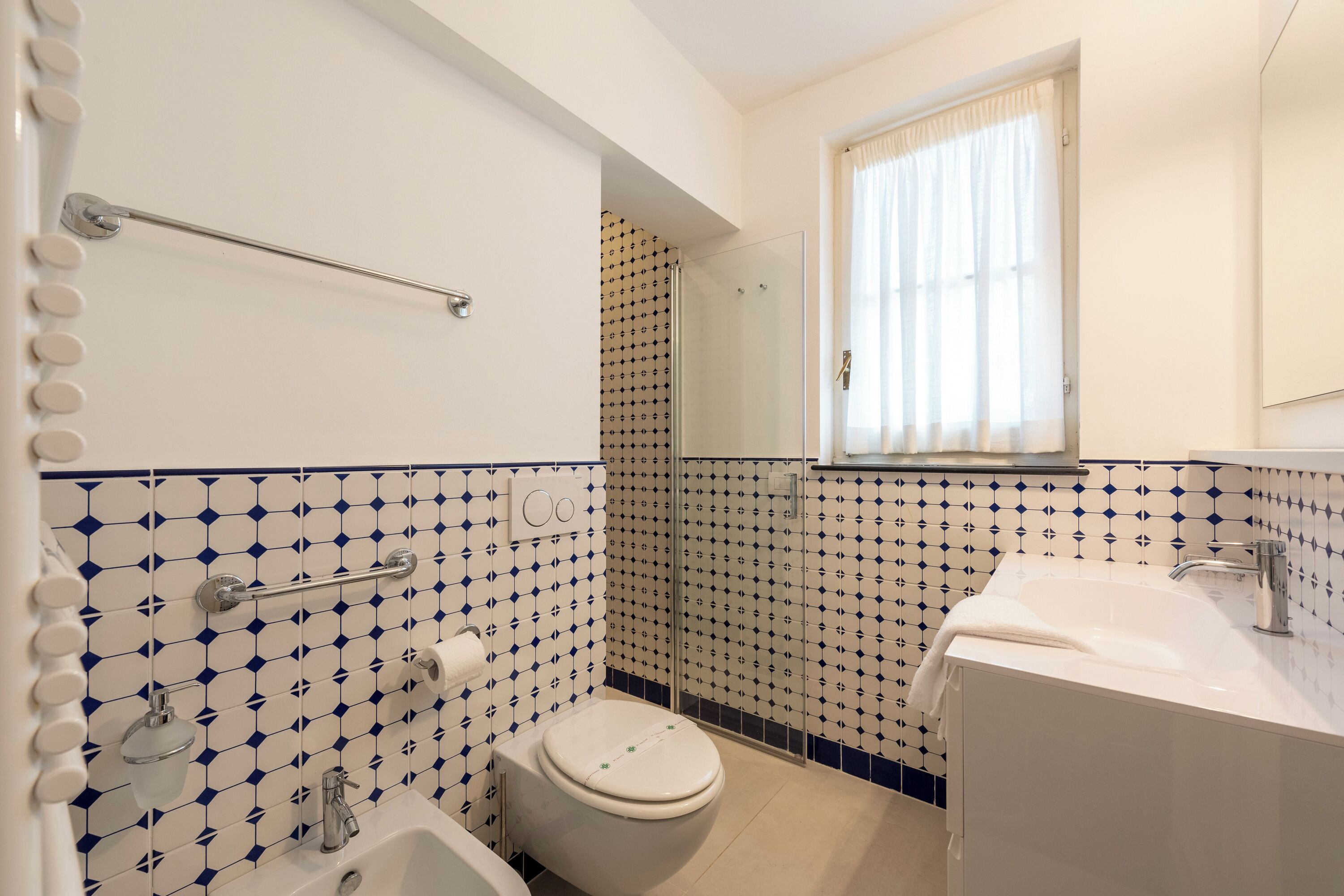 Un bagno con toilette, lavandino e doccia. Presenta un motivo a piastrelle decorate e una finestra con tende.