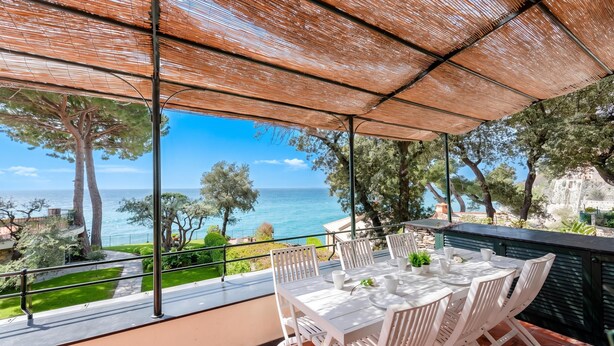 Une terrasse avec une table et des chaises, offrant une vue sur la côte, avec des arbres et un bâtiment en arrière-plan.