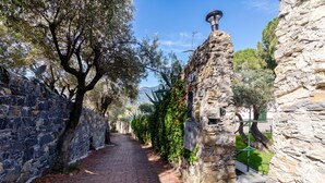 Un étroit sentier de pierres, bordé d’arbres et de murs de pierre, mène à un bâtiment en pierre surmonté d’un toit végétalisé.