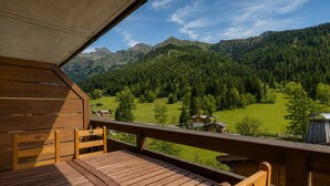 Eine Holzterrasse mit Tisch und Stühlen, mit Blick auf eine gebirgige Landschaft mit Häusern und viel Grün.