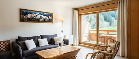 Un salon comprenant un canapé, une table basse en bois, des fauteuils en osier, un tableau et une grande fenêtre donnant sur un paysage verdoyant.
