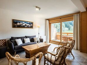 A living room with a sofa, a wooden coffee table, wicker chairs, a painting, and a large window with a view of greenery. - 3* Apartment with Wifi - 3 Bedrooms for 6 Guests in La Clusaz (La Clusaz)
