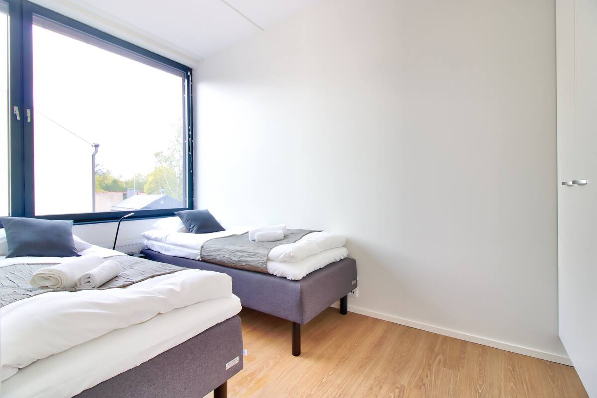 A modern hotel room with two beds, a large window, and a wooden floor.