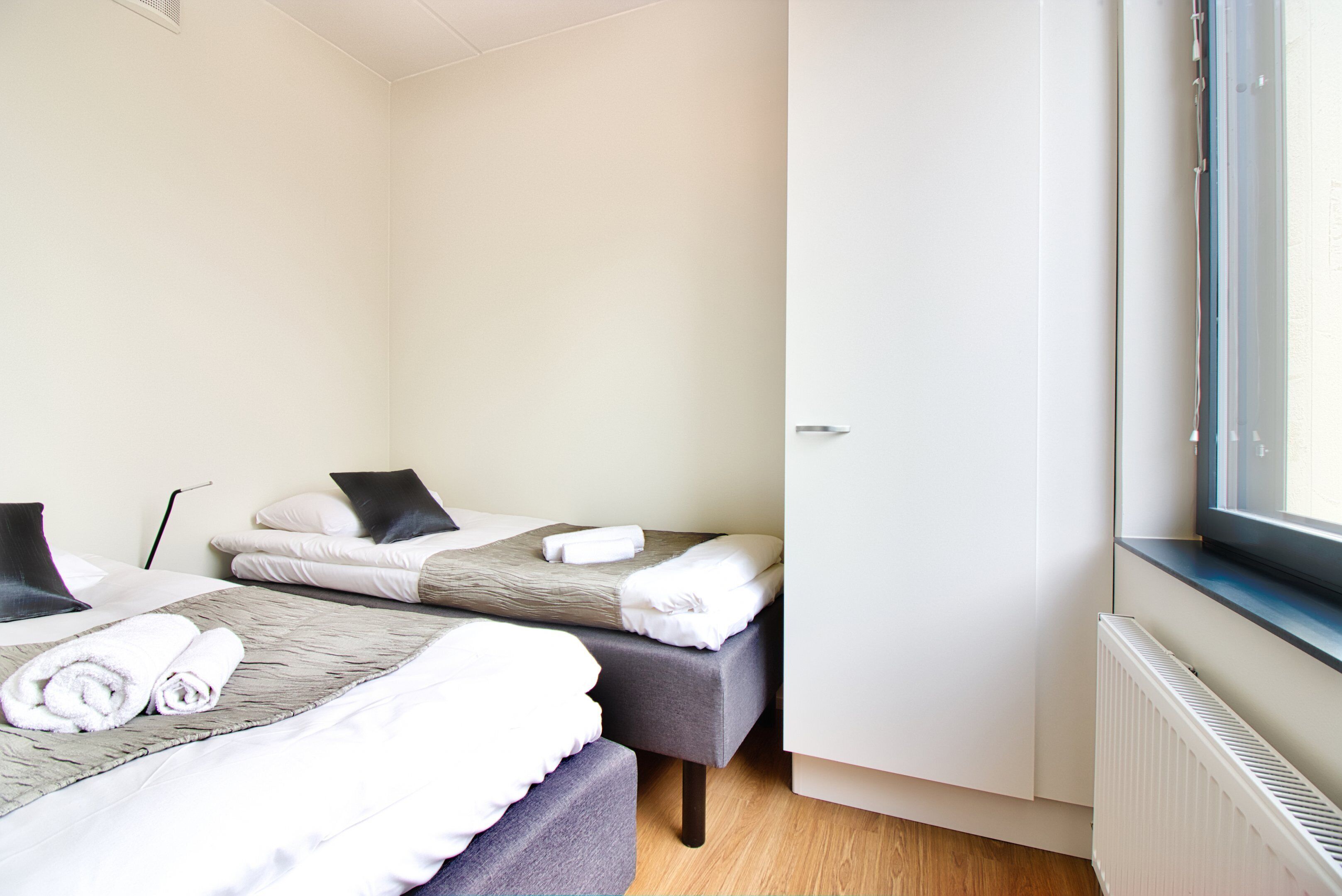 Una habitación de hotel con dos camas, cada una con cabecero gris y ropa de cama blanca, una ventana con persianas y un radiador en la pared.