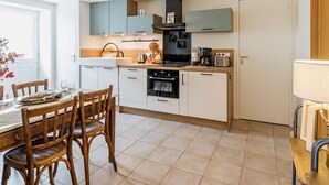 A modern kitchen with white cabinets, a black stove, and a coffee maker. There is a dining table with chairs and a window with a view. - Charming Stone Cottage with Heated Pool, Private Garden, and Close to Beaches (Saint-Lyphard)