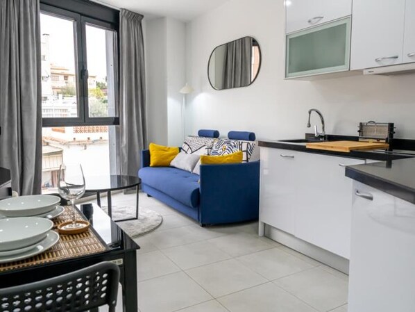 A modern apartment with a dining area, a sofa with yellow cushions, a kitchen with white cabinets, and a large window with curtains.