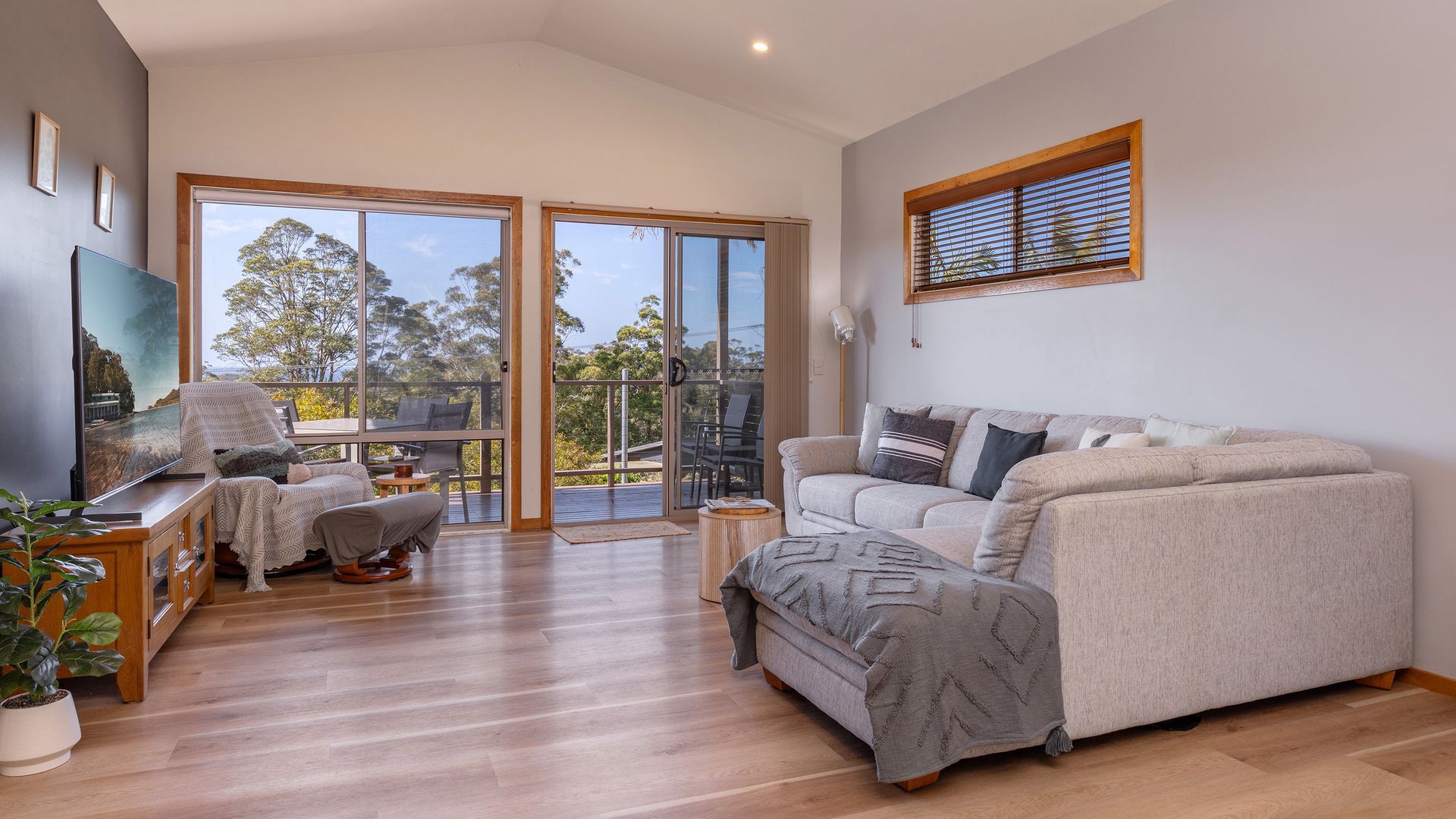 A spacious living room with a large sofa, a television, and a balcony with a view of trees.