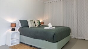 Un letto rifatto con cura, con copriletto verde, due cuscini, un asciugamano arrotolato, una lampada da comodino e un piccolo comodino bianco.