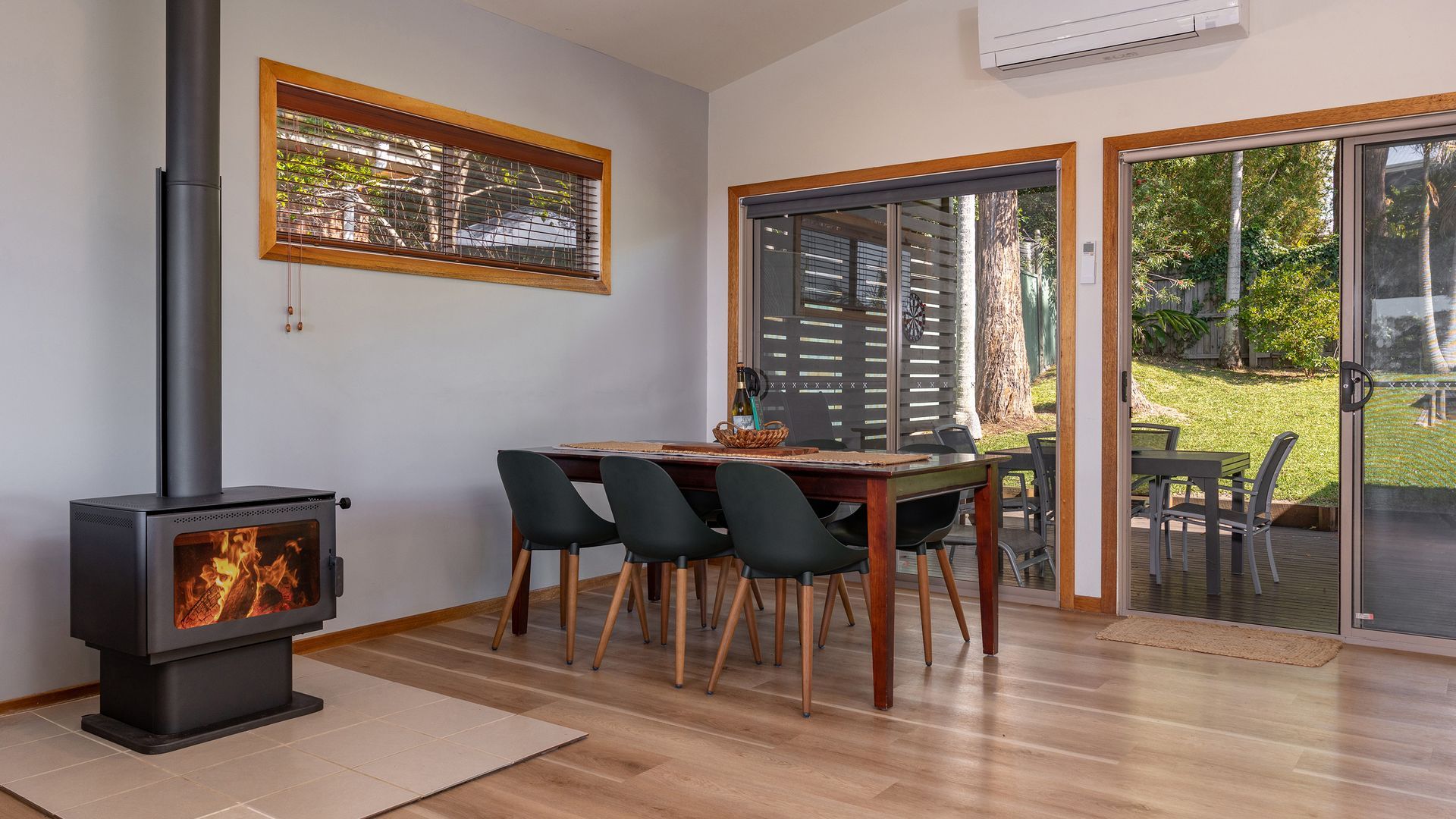 A modern living room with a wood-burning stove, a dining table with chairs, and a sliding glass door leading to an outdoor patio.