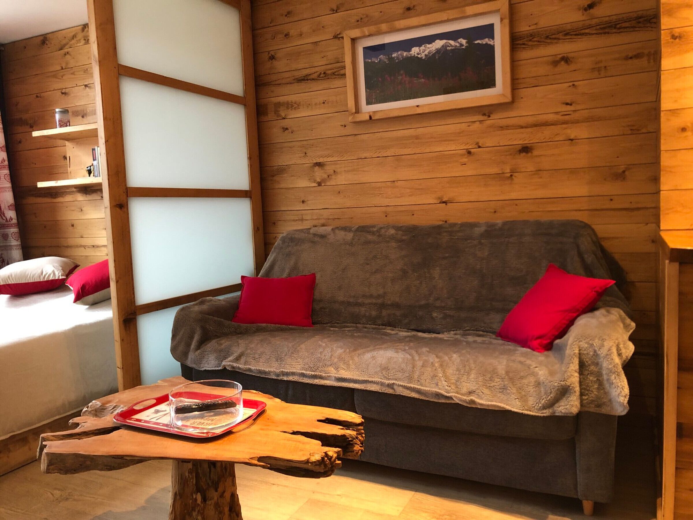Una stanza accogliente con parete in legno, un divano grigio con cuscini rossi, un tavolino in legno e un quadro incorniciato di montagne.
