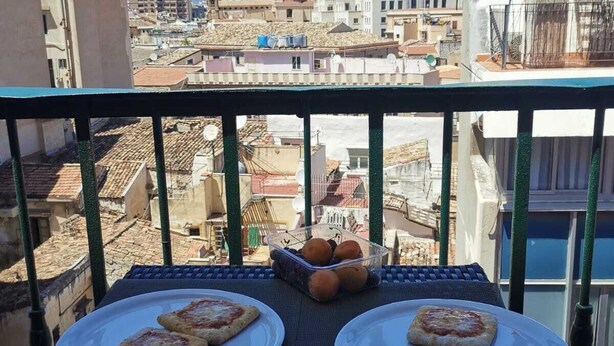 A balcony with two plates of pizza and a basket of fruit, overlooking a cityscape with buildings and a mountain. - 2 Bedroom Apartment in Palermo (Palermo)