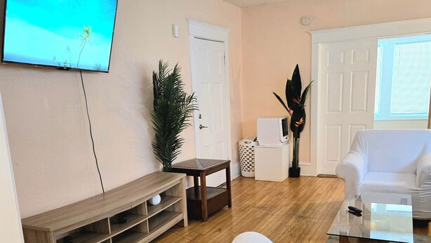 Un salon avec un téléviseur à écran plat fixé au mur, un meuble de rangement en bois, un canapé blanc, une table basse en verre et une plante en pot.