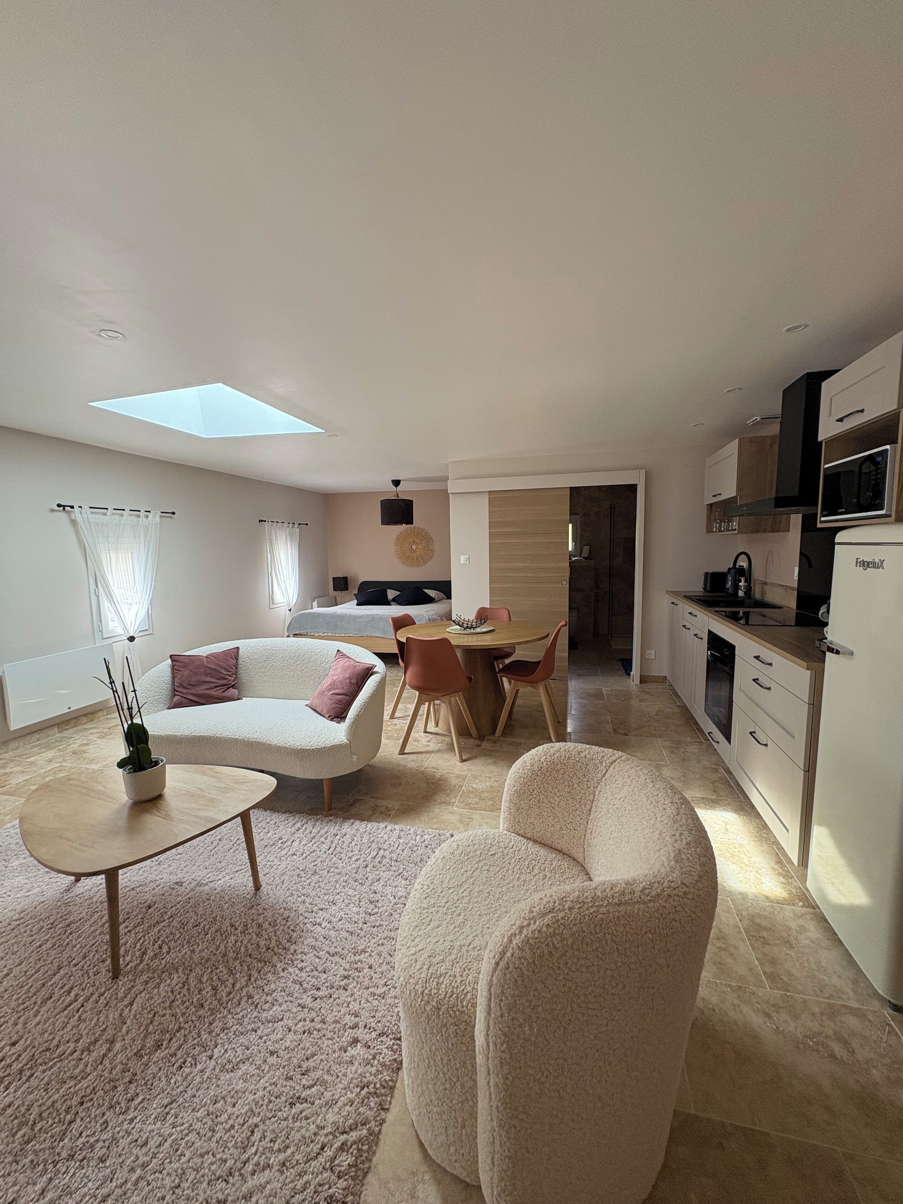 Ein modernes Wohnzimmer mit einer Couch, einem Couchtisch und einem Essbereich mit Tisch und Stühlen. Es gibt eine Küchenzeile mit einer Mikrowelle und einem Kühlschrank.