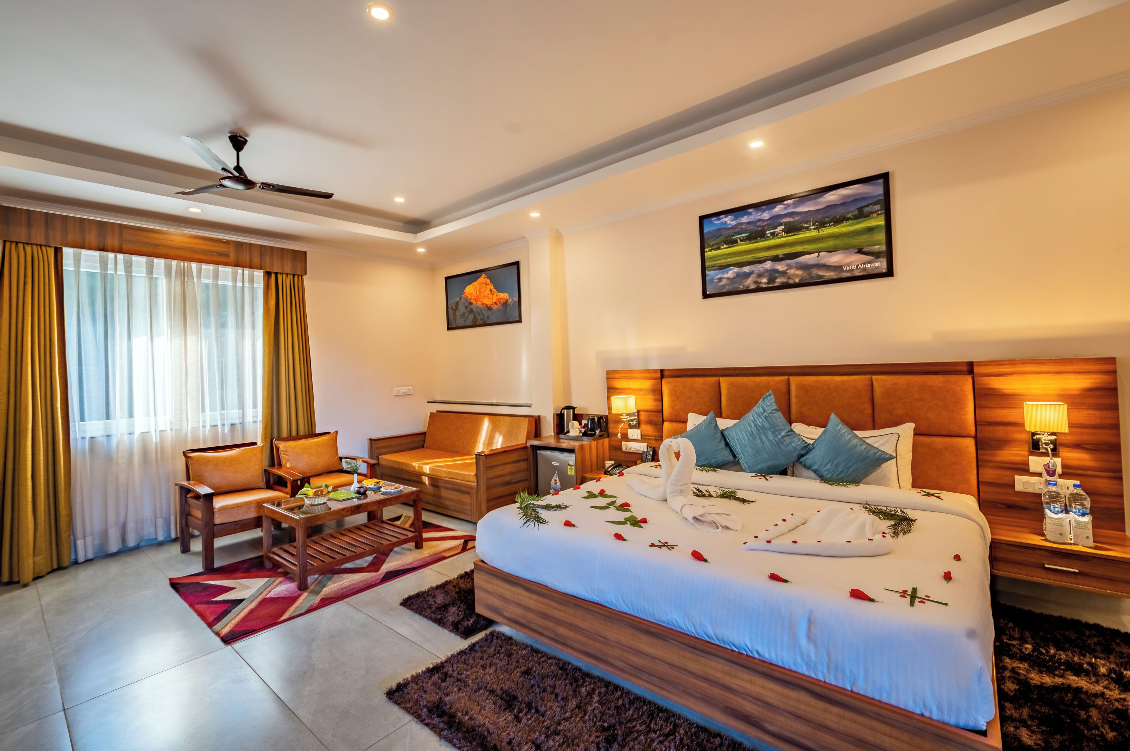Habitación de hotel con una cama grande, un escritorio, una silla y un sofá. Hay dos cuadros en la pared, un ventilador de techo y una ventana con cortinas.