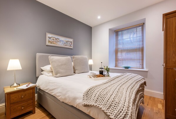 A bedroom with a bed, bedside tables, a window with blinds, a framed picture on the wall, and a wooden door.