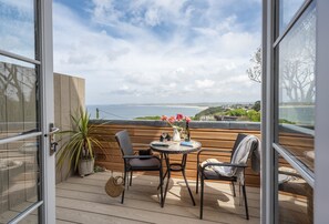 Un balcon ouvert avec un plancher en bois, deux chaises, une petite table ronde, une plante en pot et un vase rempli de fleurs.