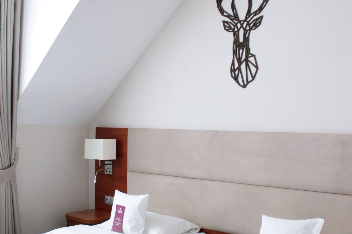 A hotel room with two beds, a wooden headboard, a wall-mounted deer head decoration, a bedside table with a lamp, and a window with curtains.
