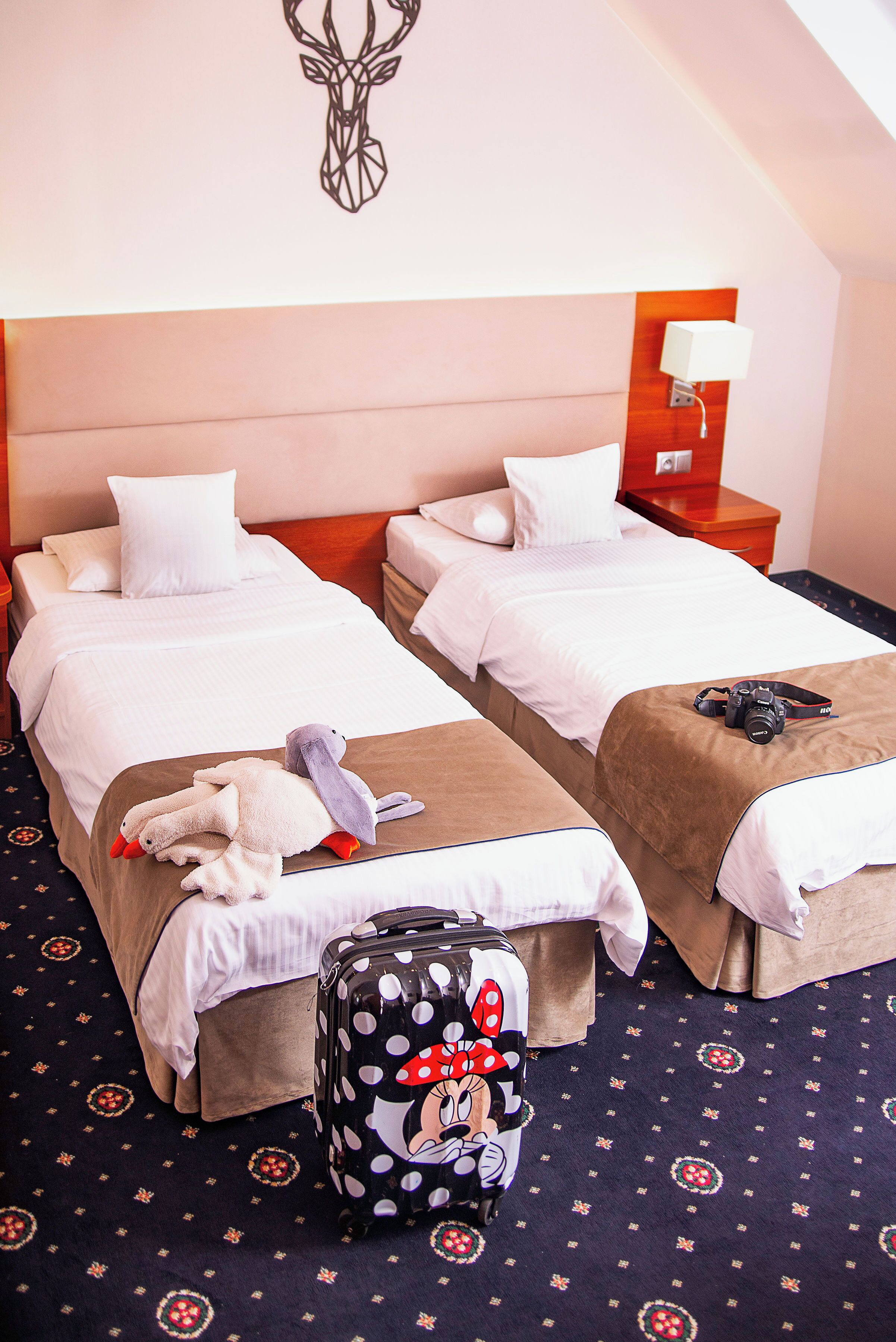 A hotel room with two beds, a nightstand with a lamp, a suitcase, and a stuffed toy on one bed.