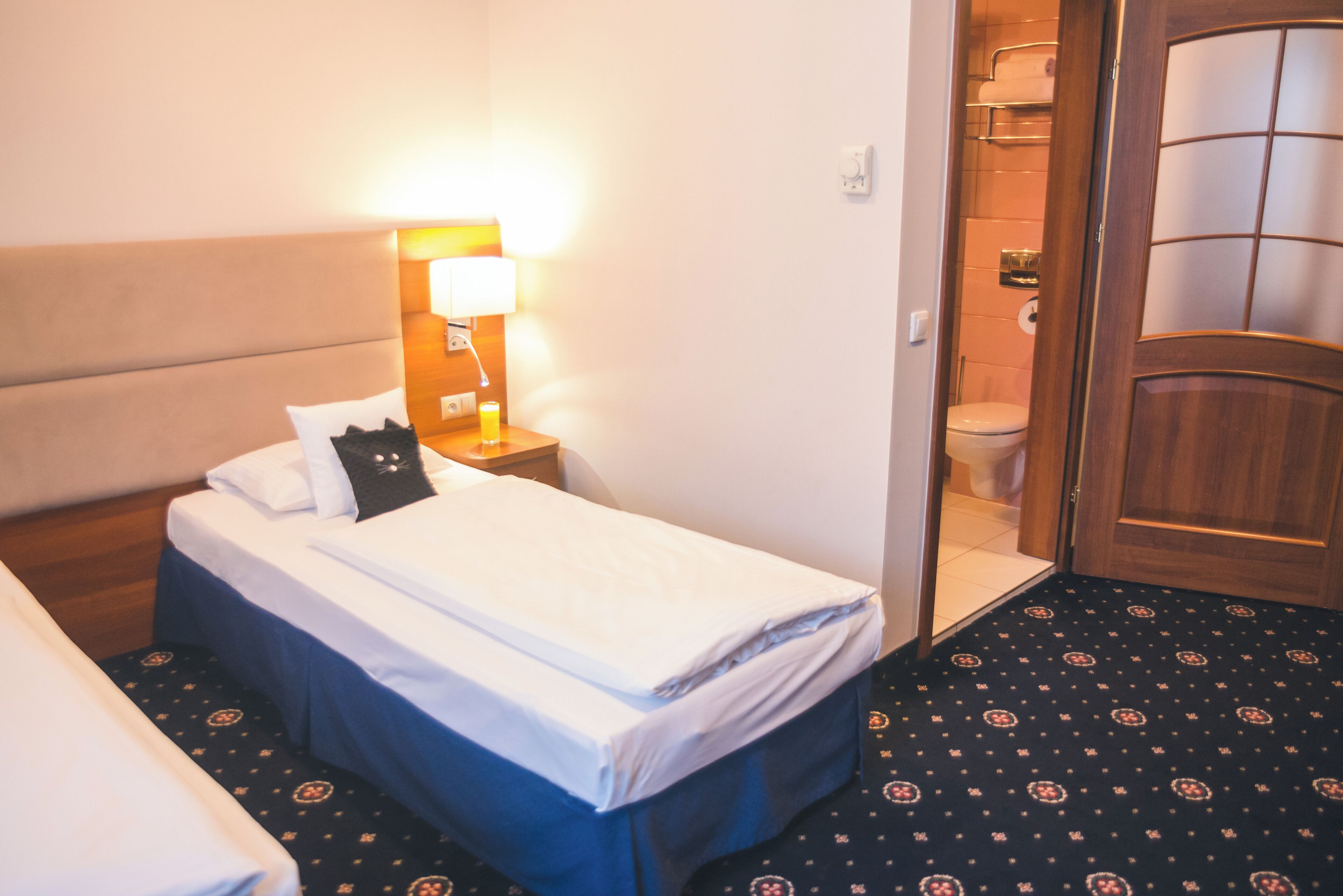 A hotel room with a bed, bedside table with a lamp, a bathroom with a toilet and shower, and a patterned carpet.