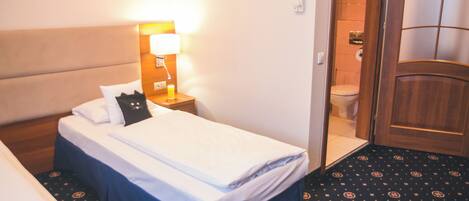 A hotel room with a bed, bedside table with a lamp, a bathroom with a toilet and shower, and a patterned carpet.