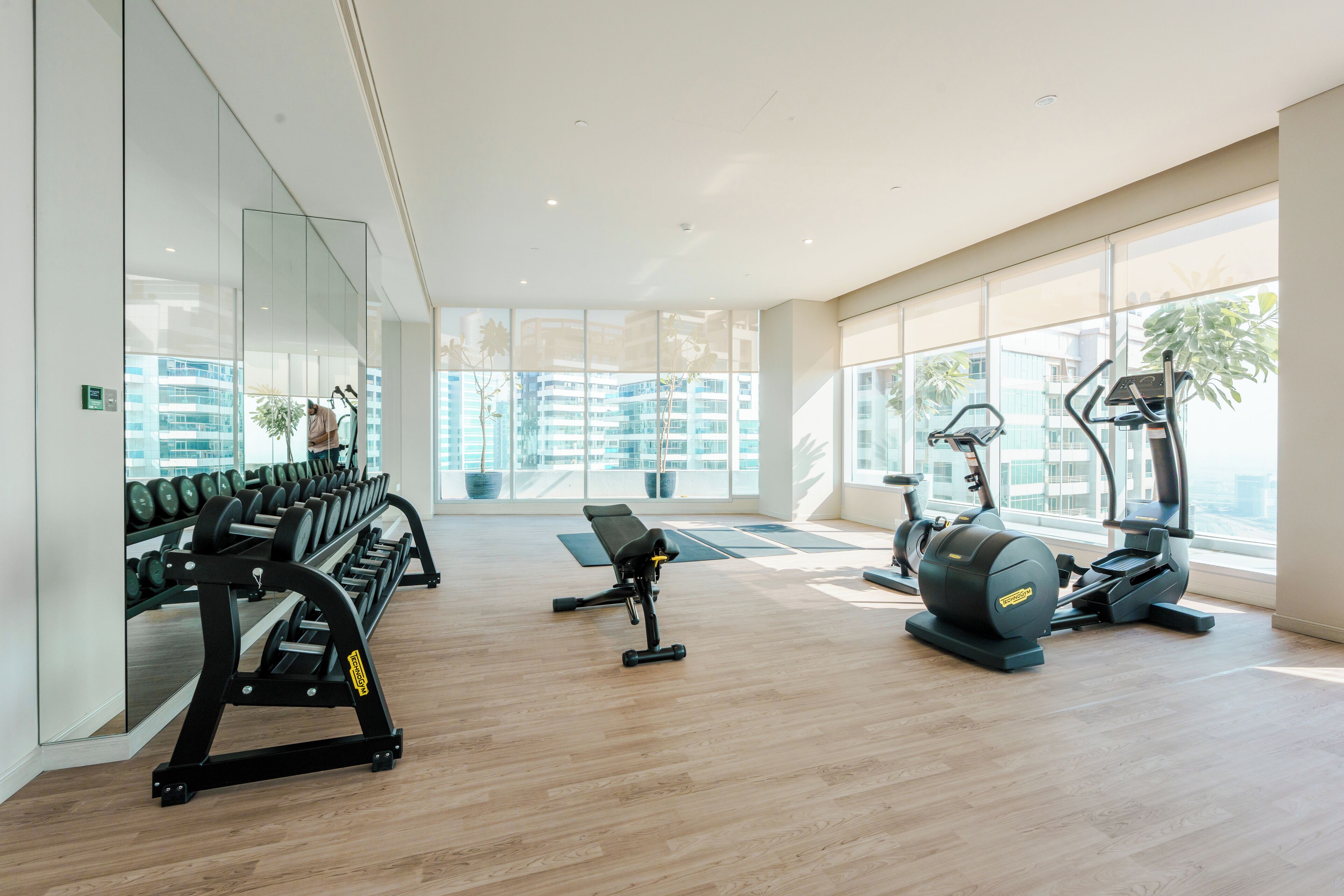 A modern gym with exercise equipment, large windows, and a wooden floor.