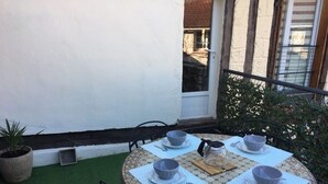 Een ronde tafel met vier grijze kommen en lepels op een tafelkleed met mozaïekpatroon, omringd door stoelen, met een potplant en een raam op de achtergrond.
