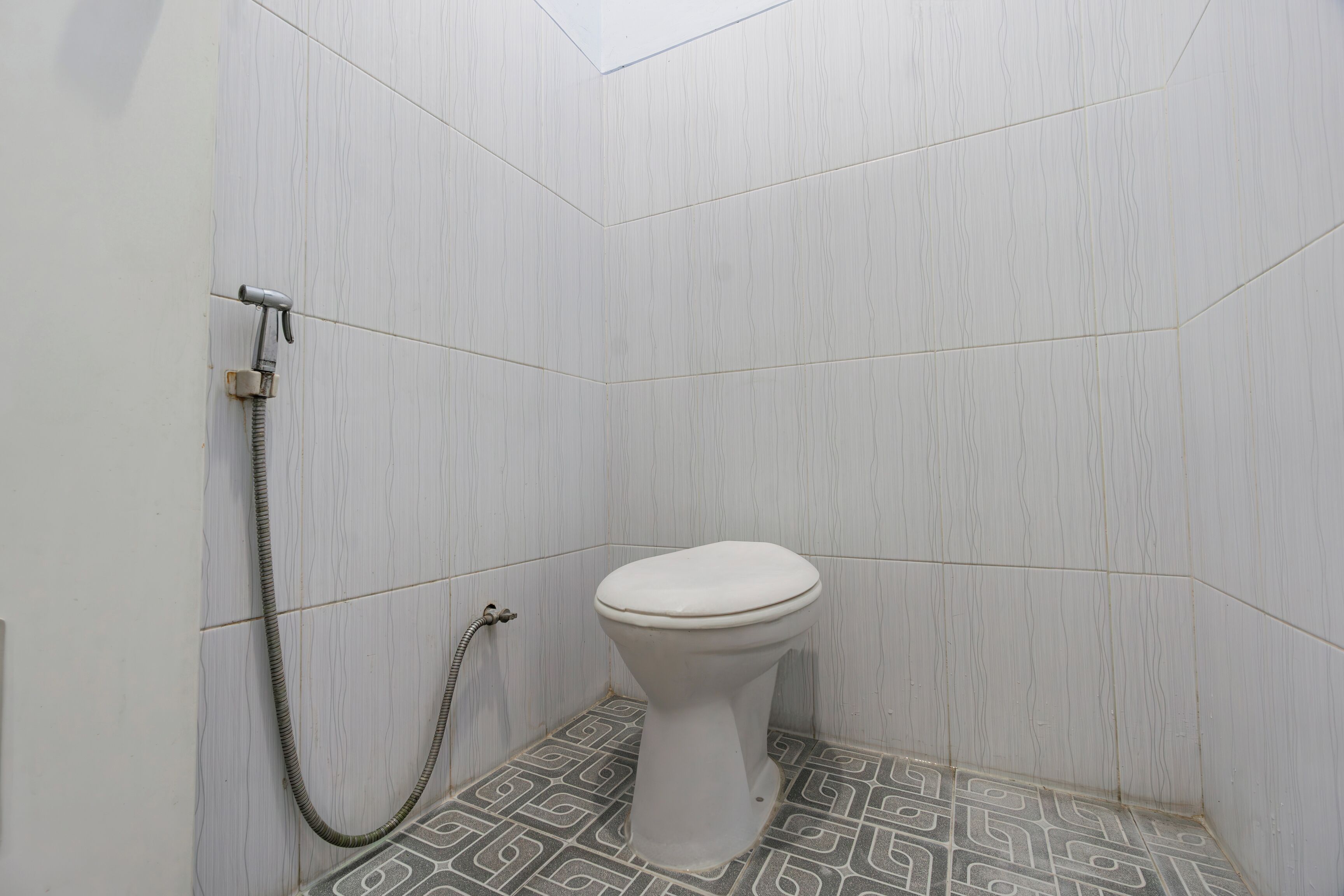Eine kleine, weiße Toilette mit Duschanschluss in einer Ecke eines Raumes mit gemusterten Fliesen.