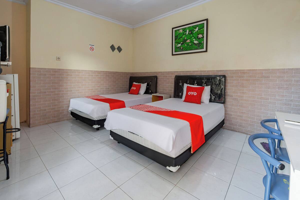 A hotel room with two beds, each with a red and white bedspread and matching pillows. The room has a tiled floor, a brick accent wall, and a painting on the wall.