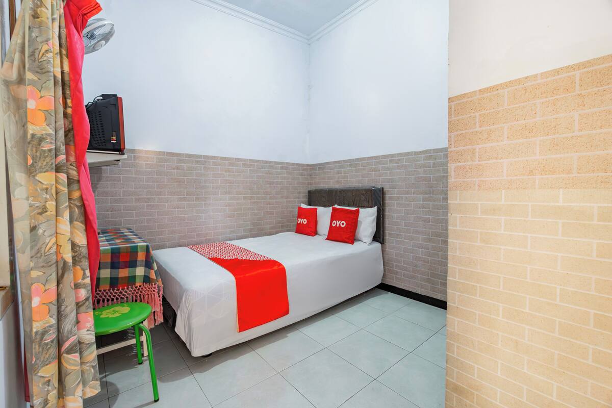 A small, single-bed room with a brick wall, a green chair, a table with a checkered tablecloth, and a red blanket on the bed.