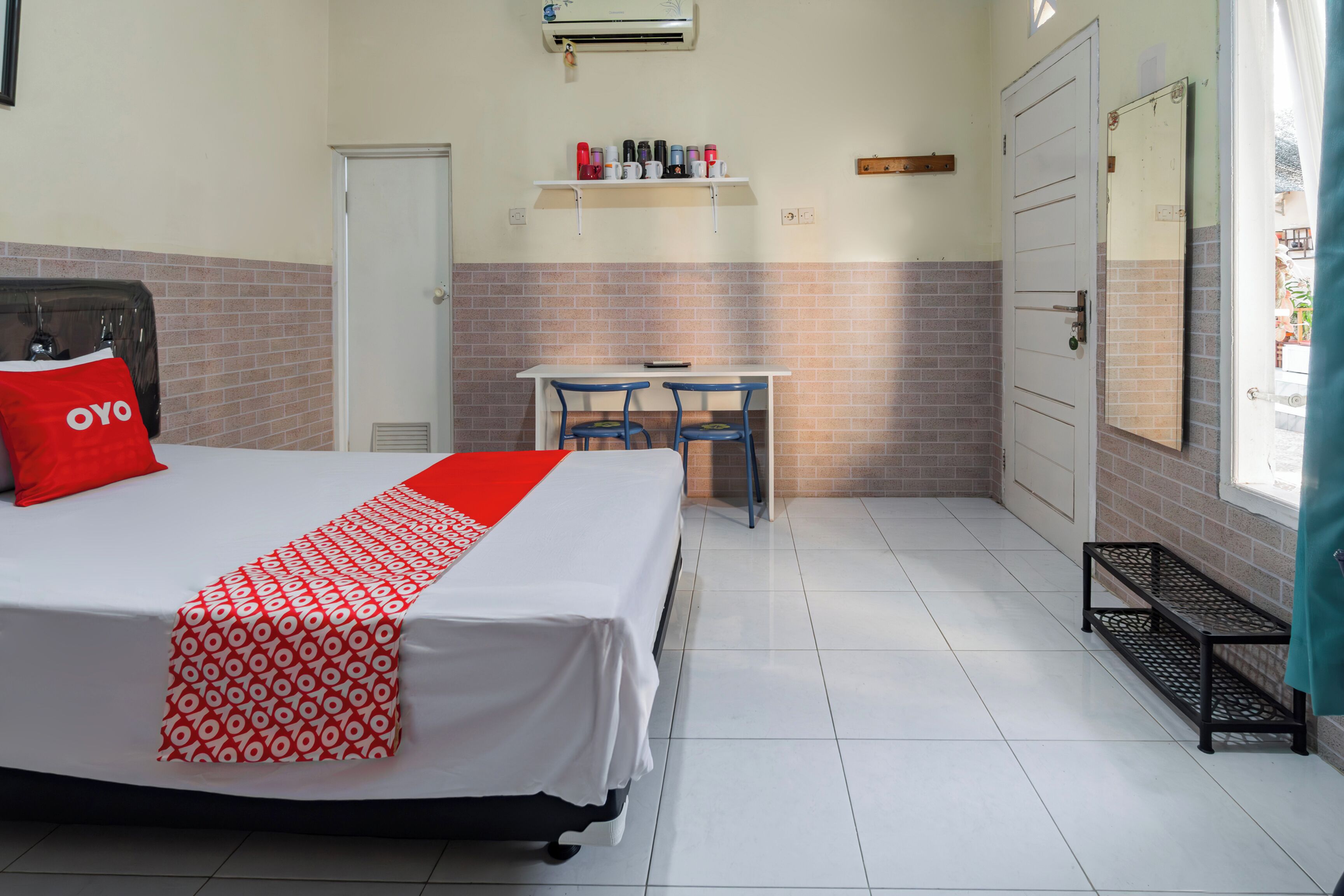 A hotel room with a bed, a desk, and chairs. There is a shelf with various items above the desk.