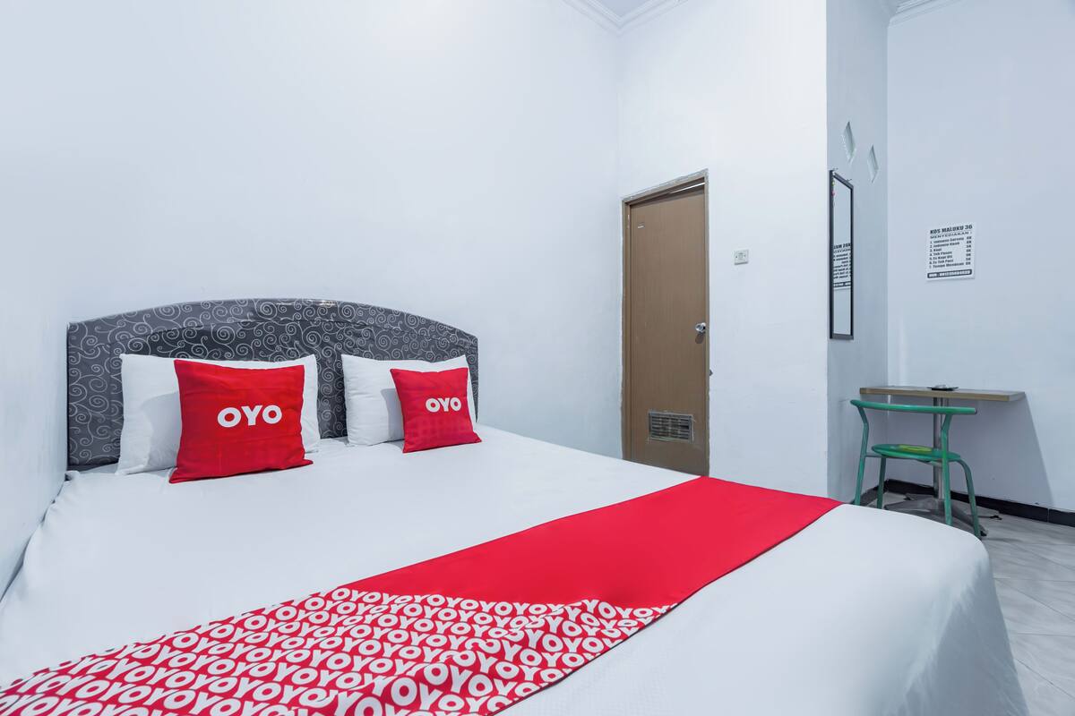 A neatly made bed with a patterned bedspread and two red pillows, a small desk with a chair, and a door with a sign on the wall.