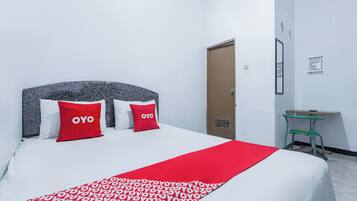 Una cama bien hecha con una colcha estampada y dos almohadas rojas, un escritorio pequeño con una silla, y una puerta con un cartel en la pared.