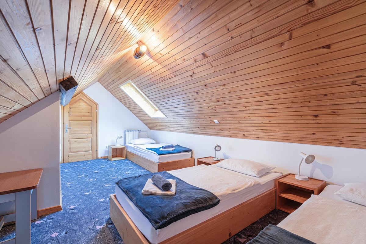 Une chambre avec un plafond en pente, des murs en bois, un puits de lumière, un lit avec un oreiller, une table de chevet et une lampe.
