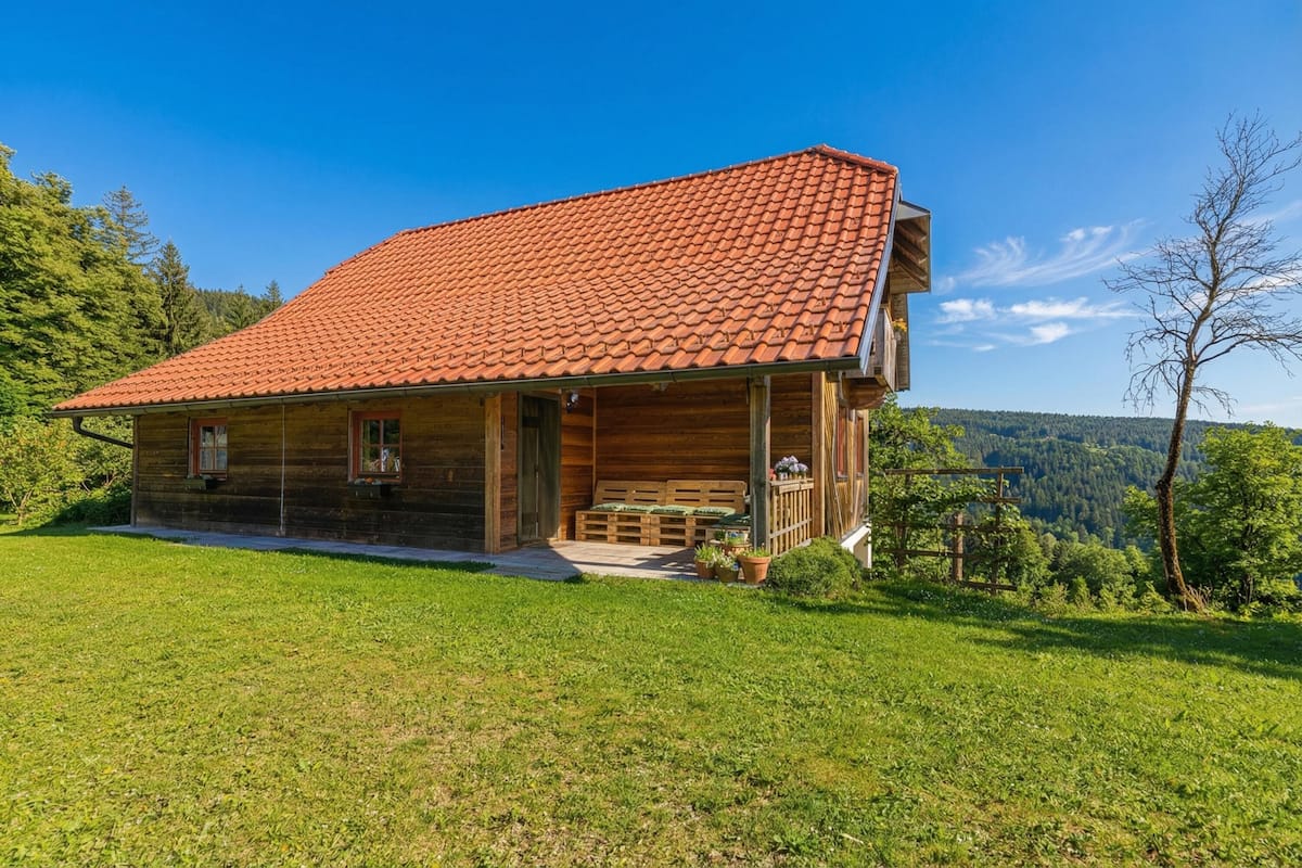 Une maison en bois avec un toit recouvert de tuiles rouges, une véranda avec des bancs et une vue sur une vallée boisée.