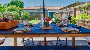 En träveranda med ett bord dukat för en måltid, omgiven av ett trästaket och ett hus med ett tegeltak i bakgrunden.