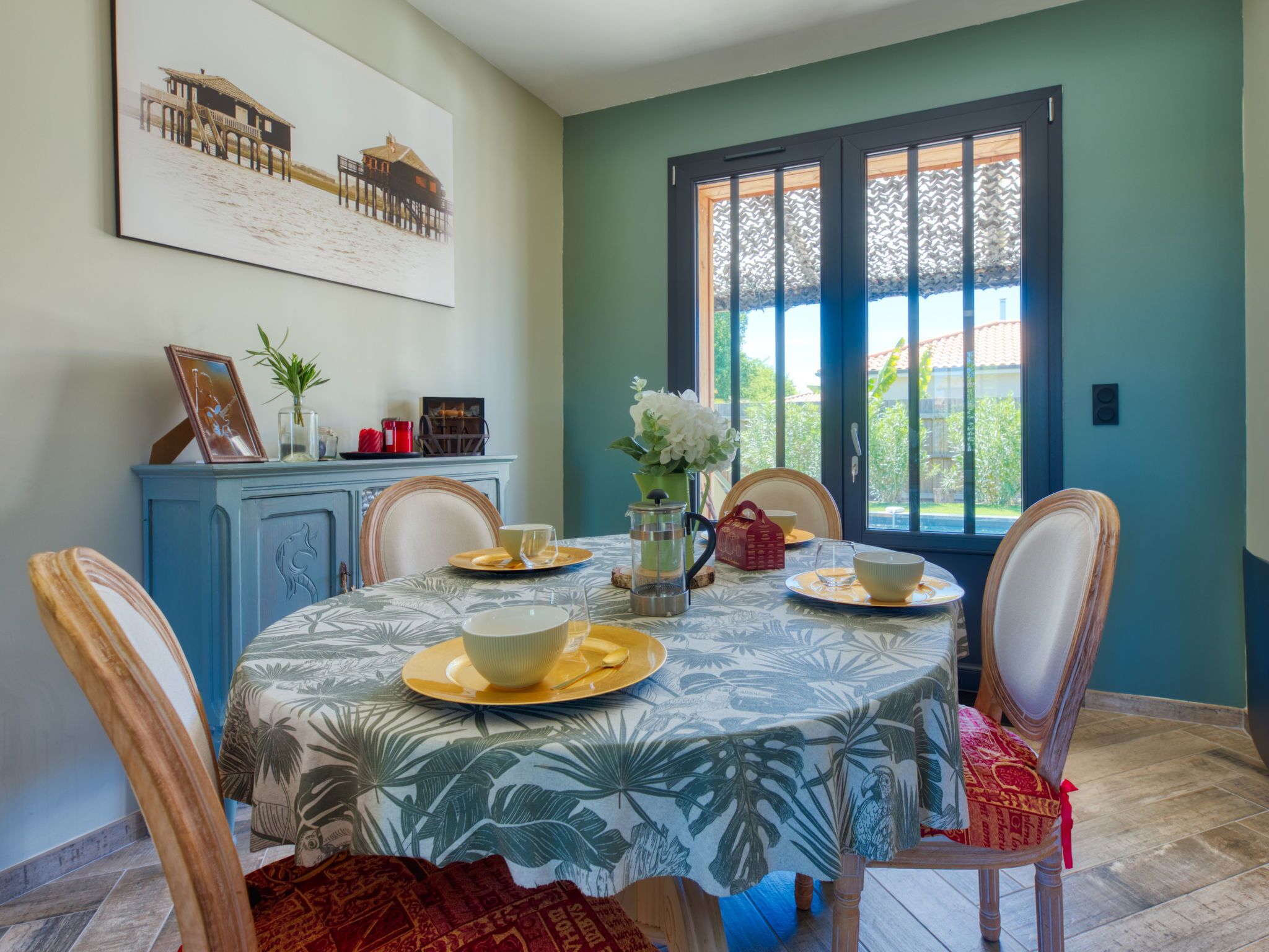 A dining area with a table set for four, a blue cabinet with decorative items, and a window with a view of greenery.