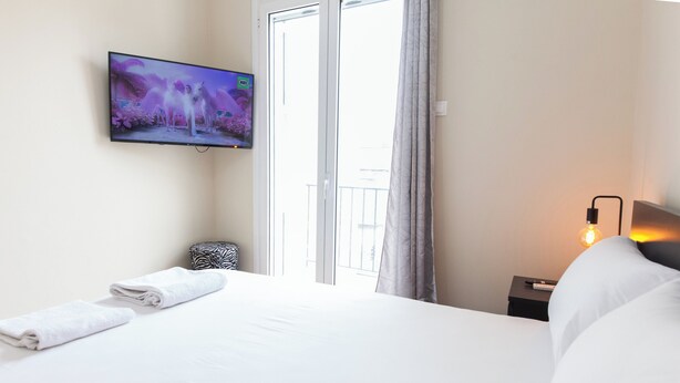 Ett hotellrum med en stor säng, en platt-TV som är monterad på väggen, ett fönster med gardiner, ett sängbord med en lampa och ett litet sidobord.